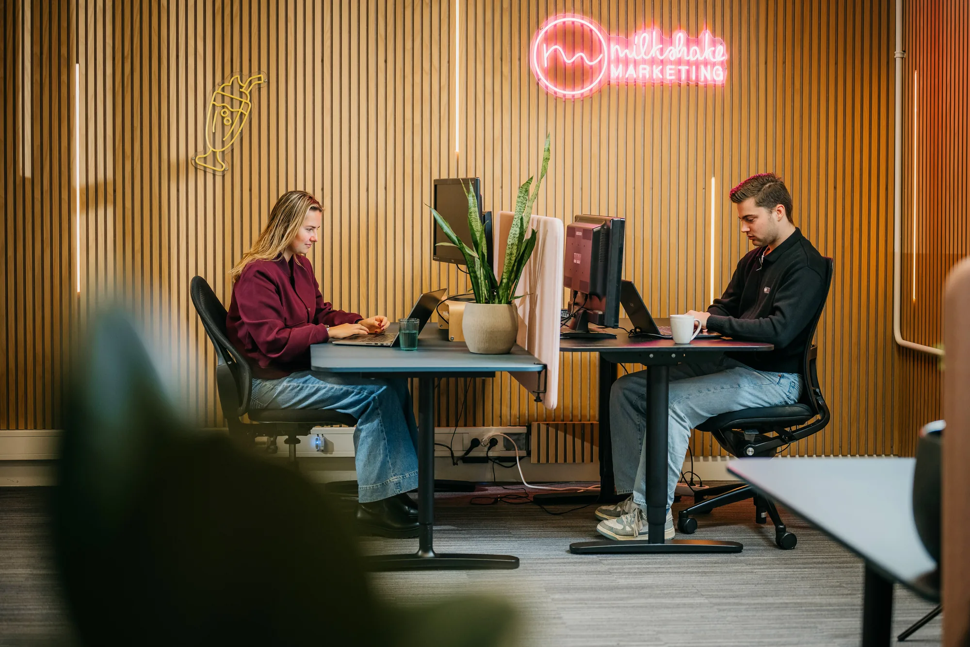 Twee collega's van Milkshake marketing werken aan hun bureau, met het neonlogo aan de houten wand achter hen